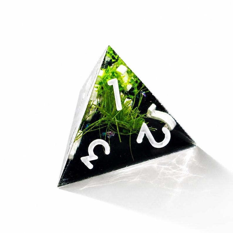 May include: A four-sided tetrahedral die made of clear resin containing green grass-like fibers and small iridescent particles. The die features white numerals 1, 2, and 3 clearly visible on the front face, set against a dark, opaque background.