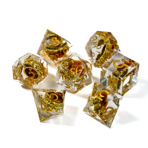 Puede incluir: Juego de siete dados de resina transparente con números dorados y diseños ornamentados en su interior. Los dados tienen varias formas geométricas, incluyendo un d4, d6, d8, d10, d12, d20 y un dado de porcentaje. Para juegos de mesa.