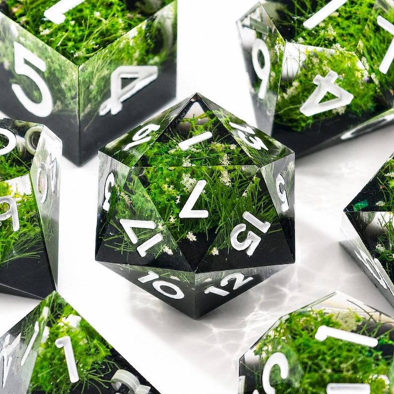 May include: A set of polyhedral dice made of clear resin, each containing embedded green moss and tiny white flowers. The dice feature sharp edges and bold, white sans-serif numerals. The base of each die is opaque black, contrasting with the clear top.