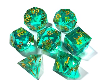 May include: Set of seven polyhedral dice in transparent teal resin. Each die features gold numbering and four-leaf clover inclusions. The dice are designed for tabletop role-playing games and are displayed on a white background.