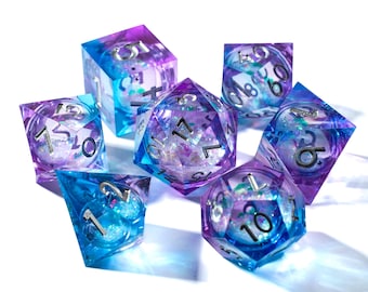 Puede incluir: Juego de siete dados poliédricos en tonos azules y morados. Los dados son transparentes con purpurina y numeración plateada. Estos dados son para juegos de rol.