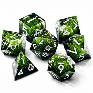 May include: A set of seven polyhedral dice made of clear resin, each containing embedded green moss and small white flowers. The dice feature sharp edges and bold, white engraved numbers, including a d20, d12, d10, d8, d6, d4, and a percentile die.