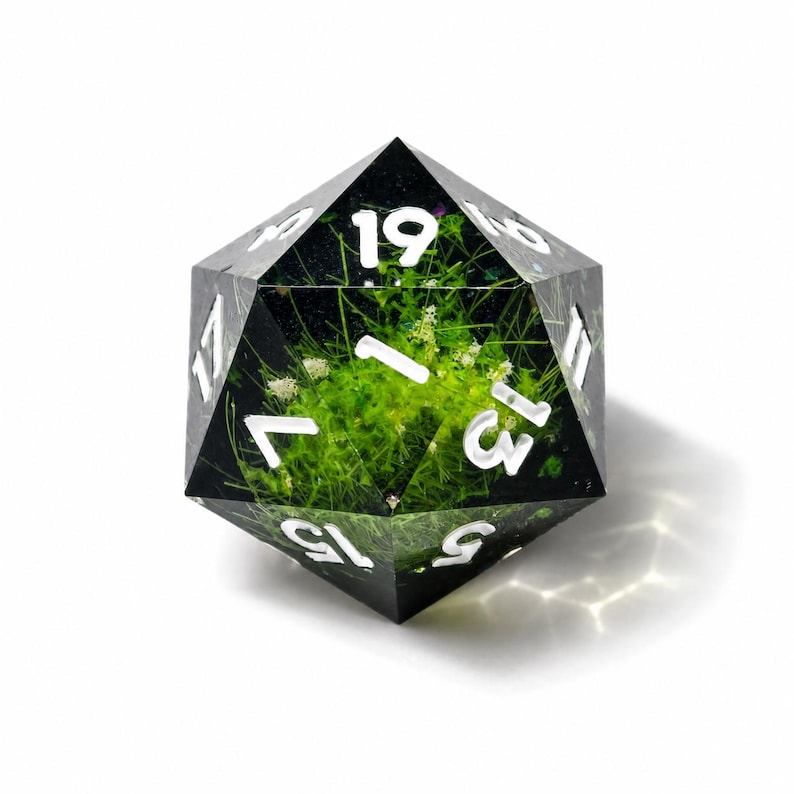 May include: A twenty-sided die made of clear resin containing embedded green grass and small white flowers. The die features sharp geometric edges and large, white, sans-serif numerals engraved into the faces, including the numbers 19, 13, 15, and 5.