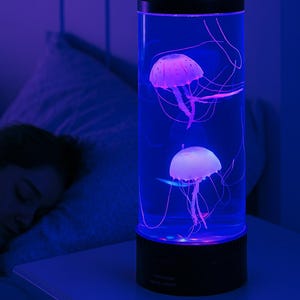Könnte beinhalten: Zylindrische Quallenlampe mit schwarzem Sockel und Deckel. Zwei Quallen schweben in dem blau beleuchteten Tank. Die Quallen sind weiß und rosa, mit langen Tentakeln. Die Lampe steht auf einem weißen Nachttisch.