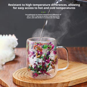 May include: A clear glass mug with a handle, filled with colorful dried flowers. The mug has the word "Valentine" printed on it. Hot water is being poured into the mug. The mug is sitting on a wooden coaster.