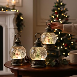 May include: Three decorative snow globe lanterns with a vintage bronze finish sit on a round wooden table. Each globe features a miniature Christmas scene with a snowman and a Christmas tree. A Christmas tree and fireplace are in the background.