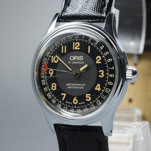May include: An Oris wristwatch with a black dial, gold-toned Arabic numerals, and a date pointer hand. The watch features a silver-toned stainless steel case, a black textured leather strap, and text reading ORIS 17 JEWELS WATERPROOF ANTISHOCK SWISS MADE.
