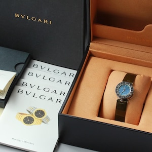 May include: A Bvlgari wristwatch with a blue dial and a dark metal band, presented in a black box with a tan interior. The set includes a black outer box, a guarantee card, and a white booklet featuring the Bvlgari logo and watch illustrations.