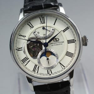 May include: A silver Orient Star automatic watch with a white face, black Roman numerals, and a black leather band. The watch features a moon phase indicator and an open heart design, showcasing the inner workings.