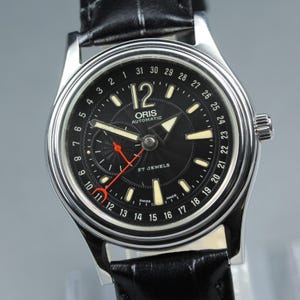 May include: An Oris Automatic wristwatch with a black dial, silver-tone case, and black leather strap. The dial features a date ring numbered 1 through 31, a red-tipped date hand, luminous hour markers, and the text Oris Automatic 27 Jewels.