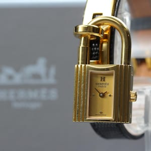 Puede incluir: Un reloj Hermès Kelly de tono dorado con una caja rectangular en forma de candado y una esfera dorada a juego. La esfera muestra el texto H HERMÈS PARIS y SWISS MADE. Está sujeto a una correa de cuero negro texturizado con hebilla dorada.