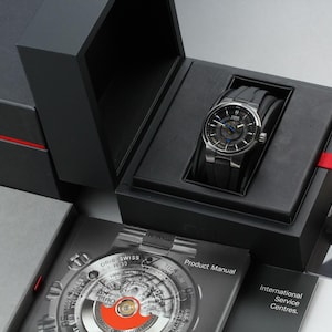 Könnte beinhalten: Eine silberfarbene Armbanduhr mit schwarzem Armband wird in einer schwarzen Box mit rotem Band präsentiert. Die Uhr hat ein dunkles Zifferblatt mit blauen Akzenten. Die Box ist geöffnet und zeigt die Uhr und eine Produktanleitung.