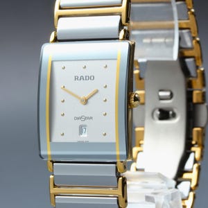 May include: A Rado DiaStar wristwatch with a rectangular silver face, gold-tone hands, and dot hour markers. The watch features a date window at the six o'clock position and a two-tone link bracelet combining silver-colored and gold-tone metal segments.