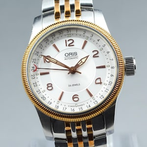 May include: An Oris Automatic wristwatch with a white dial, brown numerals, and a date pointer hand with a red crescent tip. The watch features a gold-tone fluted bezel and a two-tone stainless steel and gold-tone link bracelet with 26 jewels.