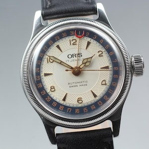 May include: A classic Oris wristwatch featuring a silver case and a black leather strap. The cream-coloured watch face has gold hands and markers, with a blue and red date ring. The watch is marked "ORIS 17 JEWELS AUTOMATIC SWISS MADE".