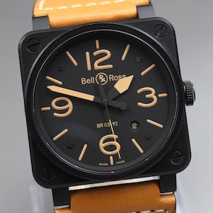 May include: A black Bell & Ross BR 03-92 watch with a square face and a tan leather strap. The watch face is black with gold numerals and hands. The watch has a screw-down crown. The words "SWISS MADE" are visible.