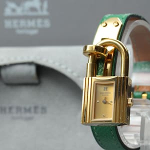May include: A gold-toned Hermes watch featuring a padlock-shaped case with a champagne-colored dial, gold hands, and the Hermes Paris logo. The watch is attached to a textured green leather strap with gold-toned hardware and a buckle closure.