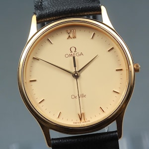 May include: An Omega De Ville wristwatch with a gold-tone round case, a cream-colored dial, and black leather strap. The dial features gold-tone hour markers, Roman numerals at the twelve and six o'clock positions, and black hands.