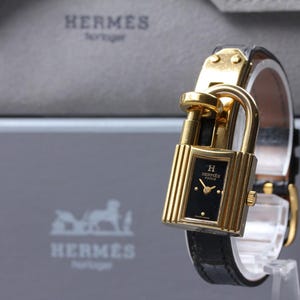 May include: A gold-toned watch with a black face and a unique padlock design. The watch features a black leather band and is displayed on a clear stand. The brand name "HERMES" is visible in the background.