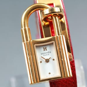 May include: A gold-tone padlock-shaped wristwatch with a white dial featuring the Hermès Paris logo and hands. The watch case is attached to a bright red textured leather strap that wraps around the top of the lock mechanism.