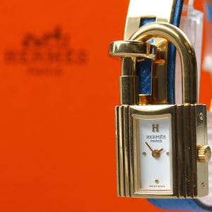 May include: A gold-tone padlock-shaped wristwatch with a white rectangular dial featuring the H logo and the text HERMES PARIS SWISS MADE. The watch has a blue leather strap and is positioned in front of an orange box with the HERMES PARIS logo.