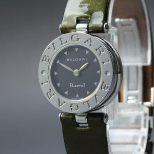 Puede incluir: Un reloj Bvlgari B.zero1 plateado con esfera gris oscuro y correa de cuero verde. La esfera del reloj presenta el logotipo de Bvlgari y el texto "B.zero1". El reloj tiene una esfera redonda con marcadores de hora plateados.