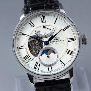 May include: A silver Orient Star automatic wristwatch with a white face, black Roman numerals, and blue hands. The watch features a moon phase indicator and a visible escapement. The black leather band complements the watch's elegant design.
