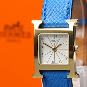 May include: A gold-tone square watch with an H-shaped case and a bright blue textured leather strap. The white dial features gold-tone hands, black Arabic numerals from 1 to 12, and the text HERMES PARIS SWISS MADE printed on the watch face.