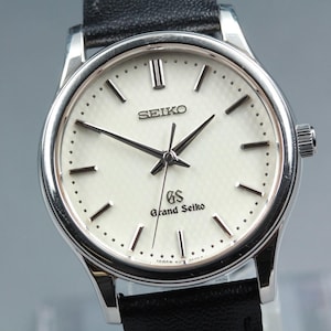 Puede incluir: Un reloj Grand Seiko plateado con esfera blanca y agujas negras. El reloj presenta el logotipo de Seiko y la inscripción "Grand Seiko". Tiene una correa de cuero negro.
