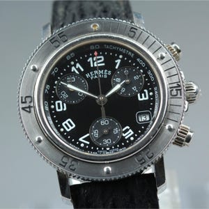 May include: A stainless steel chronograph wristwatch with a black dial, white Arabic numerals, and three sub-dials. The watch features a rotating bezel with engraved numbers, a date window at the four o'clock position, and a black textured strap.