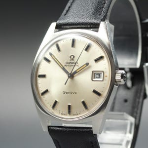 Pode incluir: Um relógio de pulso Omega Automatic Genève em tom prateado com um mostrador creme, marcadores de hora pretos, uma janela de data na posição das três horas e uma pulseira de couro preta. O mostrador exibe o logótipo e texto Omega.