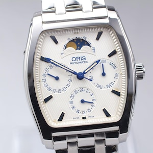 May include: A silver-toned Oris automatic watch with a white face. The watch features a moon phase indicator, day, date, and month subdials, and blue hands. The watch has a rectangular case and a stainless steel band.