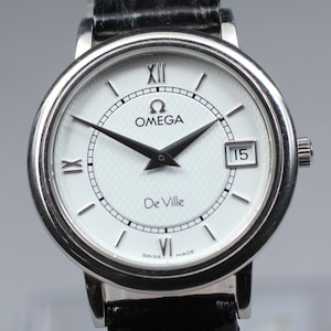 Pode incluir: Um relógio de pulso Omega De Ville prateado com mostrador branco, ponteiros pretos e numerais romanos. O relógio apresenta uma janela de data às 3 horas e uma bracelete em pele preta. O mostrador do relógio tem aproximadamente 3 cm de diâmetro.