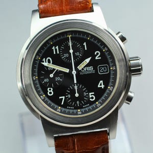 May include: A stainless steel Oris automatic watch with a black dial and a brown leather strap. The watch features a chronograph function with multiple sub-dials and a date display. The watch has a round face. The strap is brown leather.