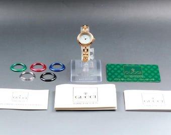 May include: A gold-toned Gucci wristwatch with a white face and a linked band. The watch is displayed on a clear stand, accompanied by colored bezels, warranty cards, and branded paperwork. The watch is a classic timepiece.