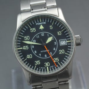 Puede incluir: Un reloj de pulsera de acero inoxidable con esfera negra. El reloj presenta números blancos, manecillas luminosas y un segundero naranja. La esfera incluye el texto "FORTIS" y "AUTOMATIC". La fecha se muestra a las 3 en punto.