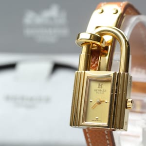 May include: A gold-tone Hermès Kelly watch featuring a rectangular case shaped like a padlock with a matching gold dial. The watch face displays the Hermès Paris logo and has gold hands. It is attached to a tan leather strap with gold hardware.