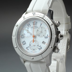 May include: A white Hermès Paris chronograph watch with a stainless steel case, a mother-of-pearl dial, and a white rubber strap. The bezel features small embedded diamonds, and the watch includes three sub-dials, a date window, and orange hands.