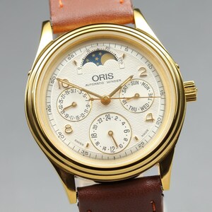 Könnte beinhalten: Eine goldfarbene Oris Automatic Winder Armbanduhr mit cremefarbenem Zifferblatt, Mondphasenanzeige, Datumsanzeige, Wochentagsanzeige und 24-Stunden-Anzeige. Die Uhr verfügt über ein braunes Lederarmband und goldfarbene Zeiger.