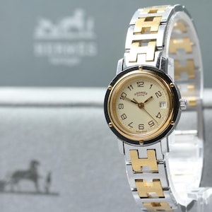 May include: A two-tone wristwatch featuring a round cream-colored dial with black Arabic numerals, the Hermes Paris logo, and a date window. The watch has a gold-tone bezel and a link bracelet alternating between silver-tone and gold-tone H-shaped links.