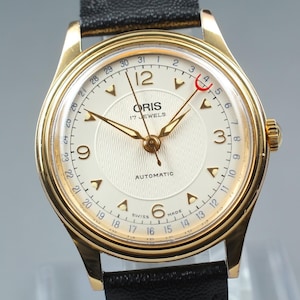 Op de afbeelding: Een goudkleurig Oris automatisch polshorloge met een crèmekleurige wijzerplaat, gouden wijzers en markeringen. Het horloge heeft een zwarte leren band en een datumweergave. De wijzerplaat leest "ORIS 17 JEWELS AUTOMATIC SWISS MADE".