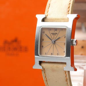 May include: A square-faced wristwatch with a silver-toned H-shaped case and a light beige leather strap. The watch dial is a matching light beige color with black Arabic numerals from 1 to 12, black hands, and the text HERMES PARIS printed in black.