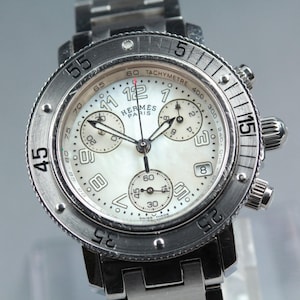 May include: A stainless steel Hermes Paris chronograph watch with a mother-of-pearl dial, featuring Arabic numerals, three sub-dials, a tachymeter scale, a date window at the four o'clock position, and a rotating bezel with engraved numbers.