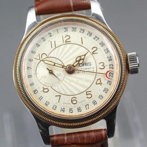 Może przedstawiać: Zegarek na rękę w stylu vintage z kremową tarczą, złotą ramką i brązowym skórzanym paskiem. Tarcza zegarka posiada czarne cyfry, małą tarczę i nazwę marki "ORIS". Zegarek posiada wskaźnik daty i teksturowane centrum.