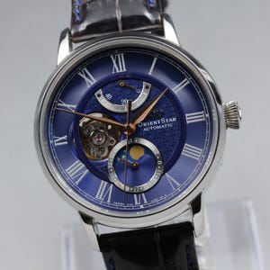 May include: An Orient Star automatic watch with a round silver-tone case and a dark blue dial. The dial features Roman numeral hour markers, a power reserve indicator, a moon phase display, and an open heart aperture. It has a dark leather strap.