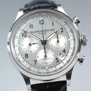 May include: A Baume & Mercier Geneve automatic chronograph watch with a silver dial, black Arabic numerals, three sub-dials, and a date window at the four o'clock position. The watch features a tachymeter scale, a telemeter scale, and a black leather strap.