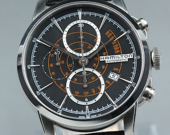 May include: A Hamilton Automatic chronograph watch with a black dial, silver-tone hands, and orange accents. The watch features a date window at the 3 o'clock position, multiple sub-dials, a tachymeter scale, and a black leather strap with orange stitching.