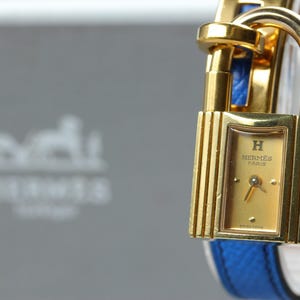 May include: A gold-toned Hermès watch with a rectangular case shaped like a padlock. The watch face is gold with the text H HERMÈS PARIS and SWISS MADE. It features a bright blue textured leather strap that loops through the top of the case.