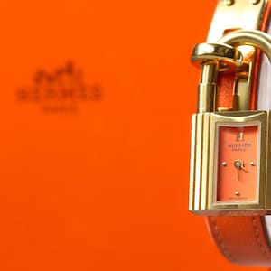 May include: A gold-toned watch with a rectangular coral-colored face and a padlock design. The watch has a brown leather strap and is set against an orange background. The watch face displays the brand name.
