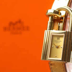 May include: A gold-toned watch with a rectangular face and a padlock-style clasp. The watch face is gold with the brand name and Swiss Made text. The watch is on a light brown leather strap, with an orange background.